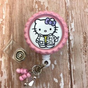 May include: Pink Hello Kitty badge reel with a white background and a Hello Kitty character wearing a white lab coat and holding a syringe. The reel has a pink and white beaded charm.