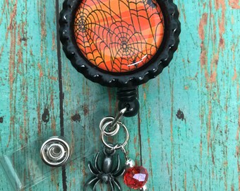 May include: A black and orange Halloween-themed retractable badge holder with a spider web design. The holder has a silver spider charm and a red crystal bead.
