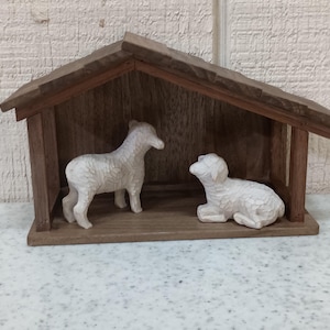 May include: A wooden nativity scene featuring a small, open-sided shelter with a sloped roof. Inside, two white carved sheep figures are positioned, one standing and one lying down. The scene is crafted from dark brown wood.