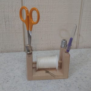 May include: A wooden sewing organizer with orange scissors, a thimble, a spool of white thread, a needle, and a purple crochet hook. The organizer is designed to hold sewing tools and supplies, keeping them organized and accessible.