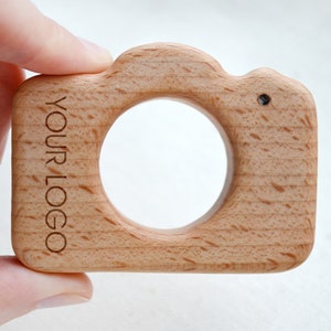 May include: A wooden camera-shaped toy with a large circular opening. The light-colored wood grain is visible. The text "YOUR LOGO" is engraved vertically on the side. A small black dot is on the top right corner.