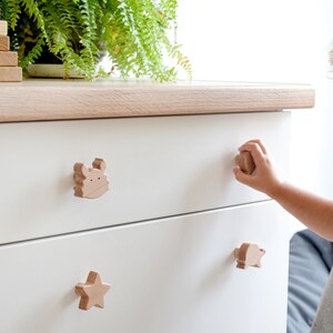 May include: White dresser with three drawers. The top drawer has a wooden crab knob, the middle drawer has a wooden star knob, and the bottom drawer has a wooden fish knob. A young child is opening the middle drawer.