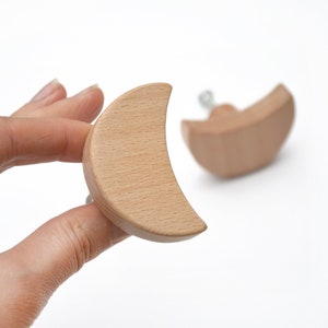 May include: Two wooden crescent-shaped cabinet knobs. The knobs are light brown and have a smooth finish. One knob is held by a hand, while the other is in the background. The knobs are designed for furniture and home decor.