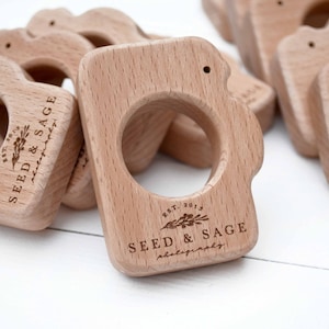 May include: A set of wooden camera-shaped teething rings with the text "EST. 2013 SEED & SAGE photography" engraved on the front.