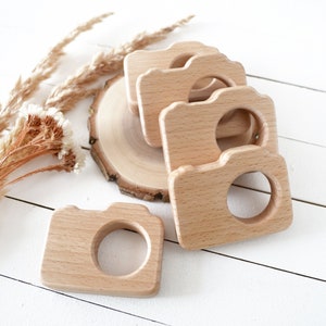 May include: Four wooden camera-shaped teethers, stacked on top of each other, on a white wooden surface. The teethers are made of natural wood and have a smooth finish.