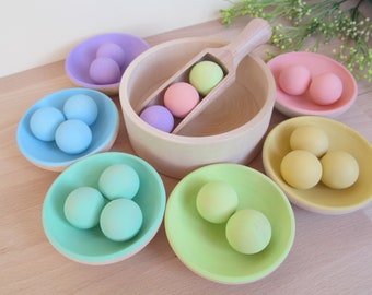 May include: A set of wooden bowls with colorful balls inside. The bowls are in shades of pink, blue, green, and yellow. The balls are also in various shades of pink, blue, green, and yellow. There is a wooden scoop with a handle and two balls inside.