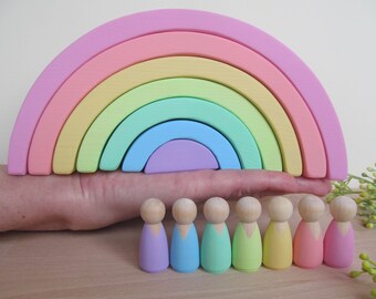 May include: A pastel rainbow wooden arch toy with seven arches in shades of pink, yellow, green, blue, and purple. Seven wooden peg dolls are lined up in front of the rainbow, each painted in a different pastel color.