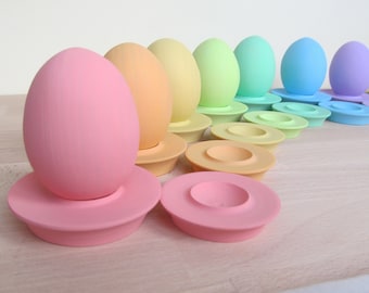 May include: A set of pastel-colored egg holders with matching eggs. The holders are arranged in a rainbow pattern, starting with pink and ending with purple. The eggs are all smooth and have a matte finish.