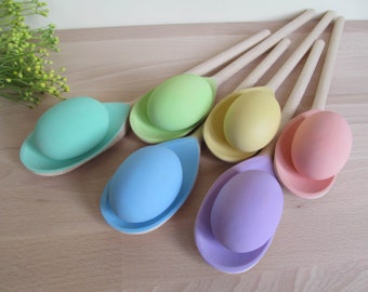 May include: Six wooden spoons with pastel colored eggs nestled inside. The spoons are arranged in a semi-circle on a light wood surface. The eggs are painted in shades of green, yellow, blue, and purple.