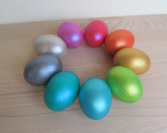 May include: Eight Easter eggs painted in different colors, including silver, gold, pink, teal, blue, green, orange, and white. The eggs are arranged in a circle on a wooden surface.