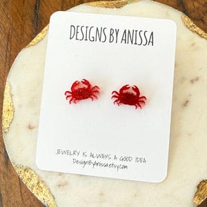 May include: Red crab stud earrings on a white card with the text "DESIGNS BY ANISSA" and "JEWELRY IS ALWAYS A GOOD IDEA DesignByAnissaetsy.com". The earrings are displayed on a decorative marble-like surface.