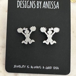 May include: A pair of silver glitter cheerleader earrings on a black card with the text "DESIGNS BY ANISSA" and "JEWELRY IS ALWAYS A GOOD IDEA".