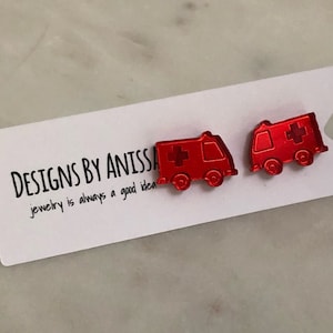 May include: A pair of red ambulance earrings with a white cross on each side. The earrings are on a white card with the text "DESIGNS BY ANISSA jewelry is always a good idea".