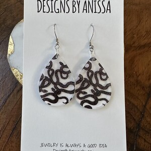 May include: A pair of silver dangle earrings with a white background and black abstract design. The earrings are on a white card with the text "DESIGNS BY ANISSA" and "JEWELRY IS ALWAYS A GOOD IDEA DesignByAnissa.etsy.com".