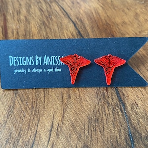 May include: A pair of red medical symbol earrings. The earrings are made of wood and have a glossy finish. The earrings are on a black card with the text "DESIGNS BY ANISSA jewelry is always a good idea".
