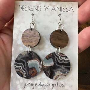 May include: A pair of earrings with a wood and agate design. The earrings are silver and have a hook closure. The wood is a dark brown color and the agate is a black, white, and brown color. The earrings are on a white card with the text "DESIGNS BY ANISSA" and "jewelry is always a good idea".