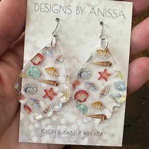 May include: A pair of clear acrylic earrings with a seashell pattern. The earrings are teardrop shaped and have silver hooks. The card behind the earrings says "Designs by Anissa" and "jewelry is always a good idea."