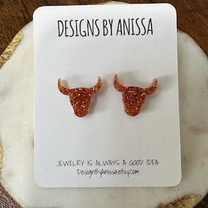 May include: A pair of orange glitter bull head stud earrings on a white card. The card reads "DESIGNS BY ANISSA" and "JEWELRY IS ALWAYS A GOOD IDEA DesignByAnissa.etsy.com". The earrings are set against a wooden background.