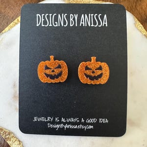 May include: A pair of orange glitter pumpkin earrings on a black card. The card reads "DESIGNS BY ANISSA" and "JEWELRY IS ALWAYS A GOOD IDEA DesignByAnissa.etsy.com". The earrings are a Halloween-themed accessory.