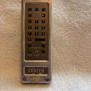 May include: Vintage Zenith TV remote control with a gold and silver metal casing. The remote features a series of buttons for volume, channel selection, and number input. The Zenith logo is visible at the bottom, along with a sticker.