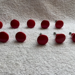 May include: A collection of ten red cabinet knobs with a glossy finish. The knobs are round with a flat top and a metal screw post on the back. They are arranged on a white, textured surface.