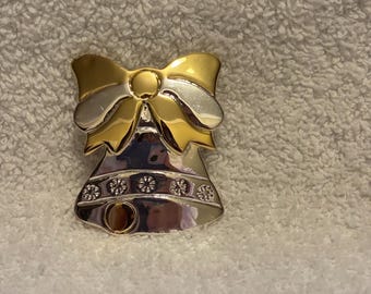 May include: A two-tone bell-shaped brooch with a gold bow and silver bell. The bell has a row of six flower-shaped embellishments. The brooch is likely made of metal and is set against a textured, off-white background.