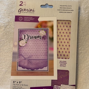 May include: A package of two Gemini embossing folders for crafting. The package features a purple and white card with the word "Dream" in white script. The package includes one embossing folder and one cut and emboss folder. The largest folder size is 5 inches by 5 inches (12.7 cm by 12.7 cm).