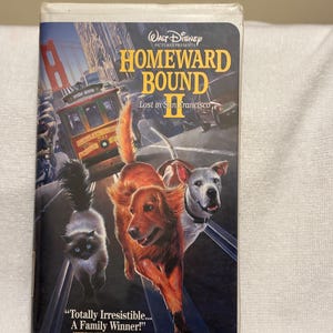 May include: A VHS tape for the Disney movie "Homeward Bound II: Lost in San Francisco." The cover features a Golden Retriever, a white dog, and a cat running. The background shows a San Francisco cable car and the Golden Gate Bridge.