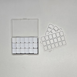 May include: A clear plastic watercolor palette with 20 white wells, open, next to a stack of blank label stickers. The palette is rectangular with a hinged lid. The stickers have a grid of 20 rectangular boxes.