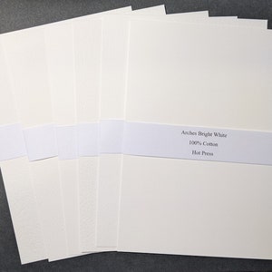 May include: A stack of bright white Arches Hot Press watercolor paper, held together with a white paper band. The band displays the text "Arches Bright White, 100% Cotton, Hot Press". The paper has a smooth texture.