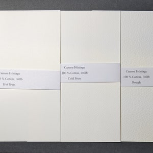 May include: Three off-white watercolor paper pads with varying textures. Each pad has a white label with the text "Canson Heritage 100% Cotton, 140lb" and a different finish: Hot Press, Cold Press, and Rough. The paper is on a dark gray surface.