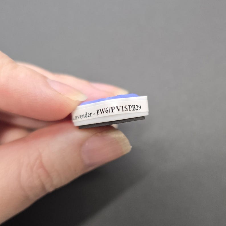 May include: A small, rectangular magnet with a white border and a lavender top. The text "Lavender - PW6/PV15/PB29" is printed on the side. The magnet is held between fingers against a grey background.