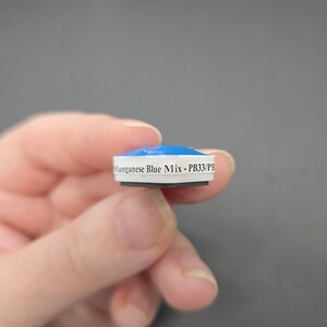 May include: A small, round object with a blue top and white sides. The text "Manganese Blue Mix - PB33/PB" is printed on the side. The object is held in a person's hand against a gray background.