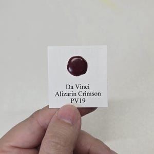 May include: A small white card with the text "Da Vinci Alizarin Crimson PV19" and a dark red paint swatch. The paint swatch is a circular shape. The card is held by a person.