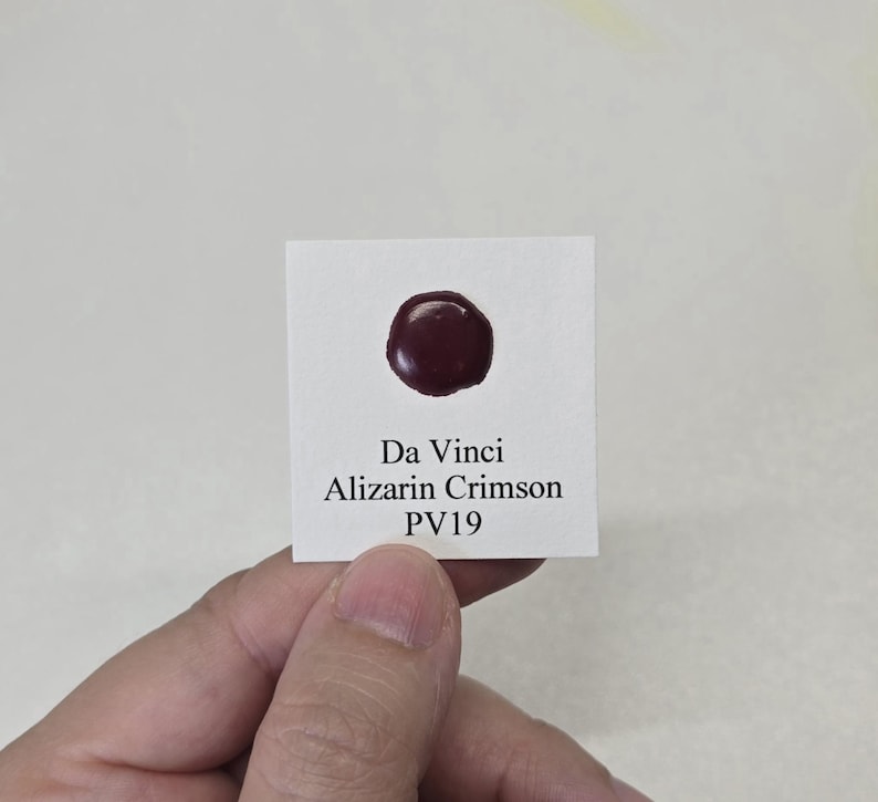 May include: A small white card with the text "Da Vinci Alizarin Crimson PV19" and a dark red paint swatch. The paint swatch is a circular shape. The card is held by a person.