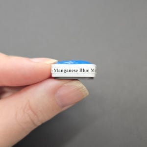 May include: A small, rectangular magnet with a blue top and white sides. The text "-Manganese Blue Mi" is printed on the side. The magnet is held between fingers against a gray background.