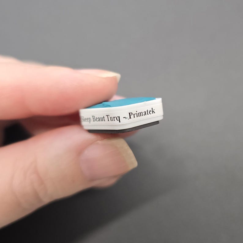 May include: A small, rectangular object with a light blue top and white sides. The text "Sleep Beaut Turq ~ Primatek" is printed on the side. The object is held in a person's hand.