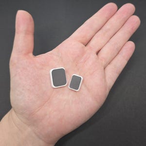 May include: Two square-shaped, gray and white objects are displayed in an open hand. The objects have a dark gray center and a white border. The larger square is slightly bigger than the smaller one. The background is a solid, neutral color.