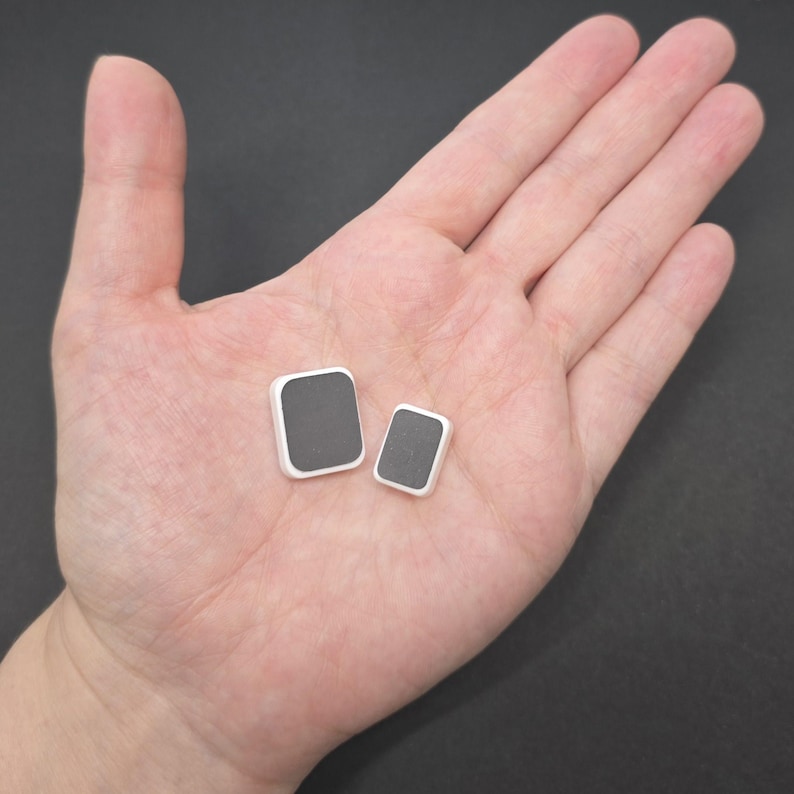 May include: Two square-shaped, gray and white objects are displayed in an open hand. The objects have a dark gray center and a white border. The larger square is slightly bigger than the smaller one. The background is a solid, neutral color.