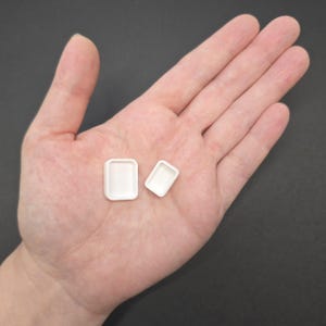 May include: Two small, white, rectangular trays. One tray is slightly larger than the other. The trays are empty and have rounded corners. The image is taken from a top-down perspective, with the trays resting on a person's open palm.