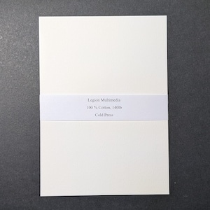 May include: A stack of off-white Legion Multimedia cold press paper. The paper is held together by a white band with the text "Legion Multimedia 100% Cotton, 140lb Cold Press". The paper is on a dark gray surface.