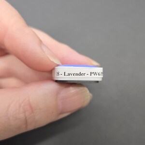 May include: A small, rectangular object with a light blue top and white sides is held between fingers. The text "DS - Lavender - PW6/P" is printed on the side. The object appears to be a small product sample or component.