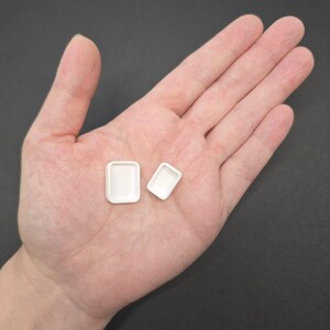 May include: Two small, white, rectangular trays. One tray is slightly larger than the other. The trays are empty and have rounded corners. The image is taken from a top-down perspective, with the trays resting on a person's open palm.