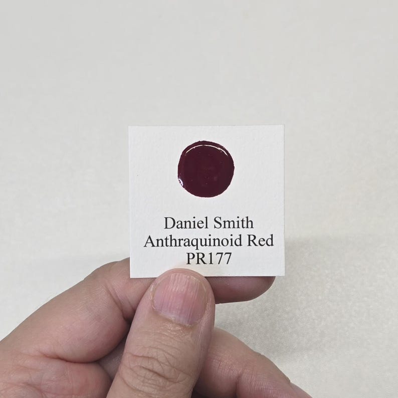 May include: A small white card with a dark red circular paint swatch. The card has the text "Daniel Smith Anthraquinoid Red PR177" printed in black. The card is held by a person.