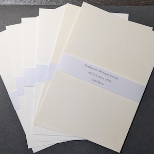 May include: A stack of cream-colored Strathmore 500 Series Gemini cold press paper. The paper is 100% cotton and 140lb. A white band with the product details is wrapped around the stack. The paper has a textured surface and is arranged on a dark surface.