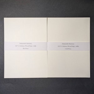 May include: Two off-white paper pads with text reading "Hahnemühle Harmony 100% Cellulose (Wood Pulp), 140lb Hot Press" and "Hahnemühle Harmony 100% Cellulose (Wood Pulp), 140lb Cold Press". The pads are on a dark gray surface.