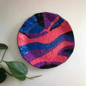 May include: A round, colorful decorative plate with a textured surface. The plate features a vibrant design of pink, blue, purple, and black wavy stripes. The plate is a unique piece of art.