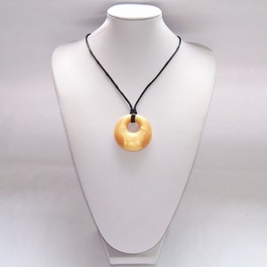 May include: A pendant necklace featuring a circular, donut-shaped pendant in a light tan color. The pendant is suspended from a black cord. The necklace is displayed on a white mannequin.