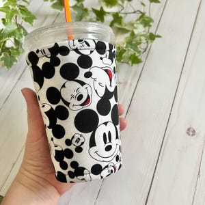 May include: A white and black fabric drink sleeve featuring a pattern of Mickey Mouse faces and black polka dots. The sleeve is wrapped around a clear plastic cup with a lid and an orange straw. The design is a fun and playful accessory.