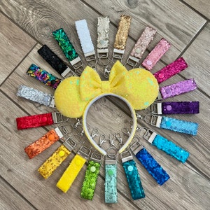 May include: A set of 18 colorful sequin keychains with silver key rings. The keychains are arranged in a circle around a yellow sequin headband with a bow.